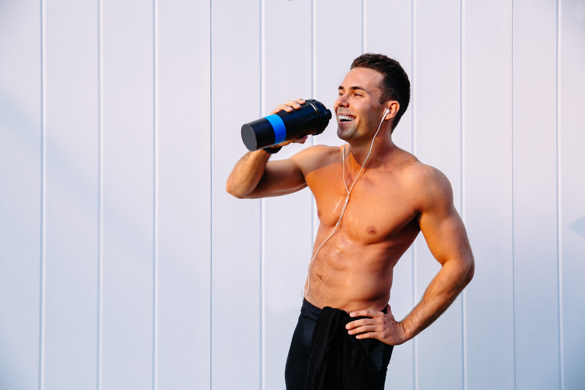 Joyful muscular guy enjoying drinking a water after workout, listening to music in earphones. Standing against the white wall. Sport concept.